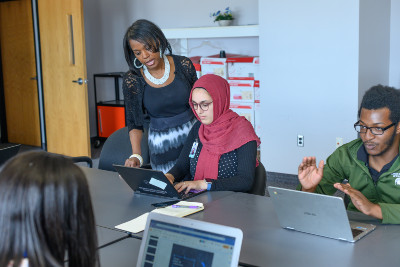 Black woman standing over the shoulder of a seated student wearing a hijab. Both are looking at a laptop.