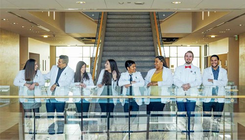 A group of people in white lab coats stand behind a glass railing and converse with each other.